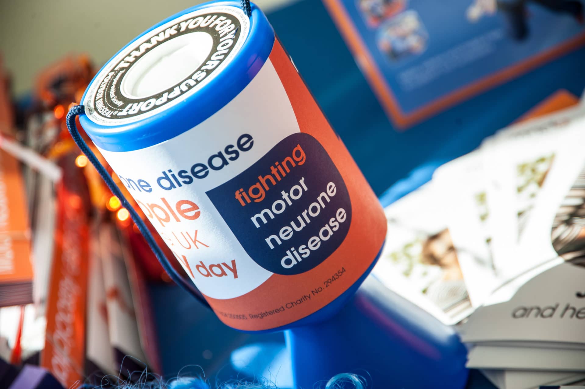 Blue, whit and orange MND Association collection tin sat on a table with MND related literature