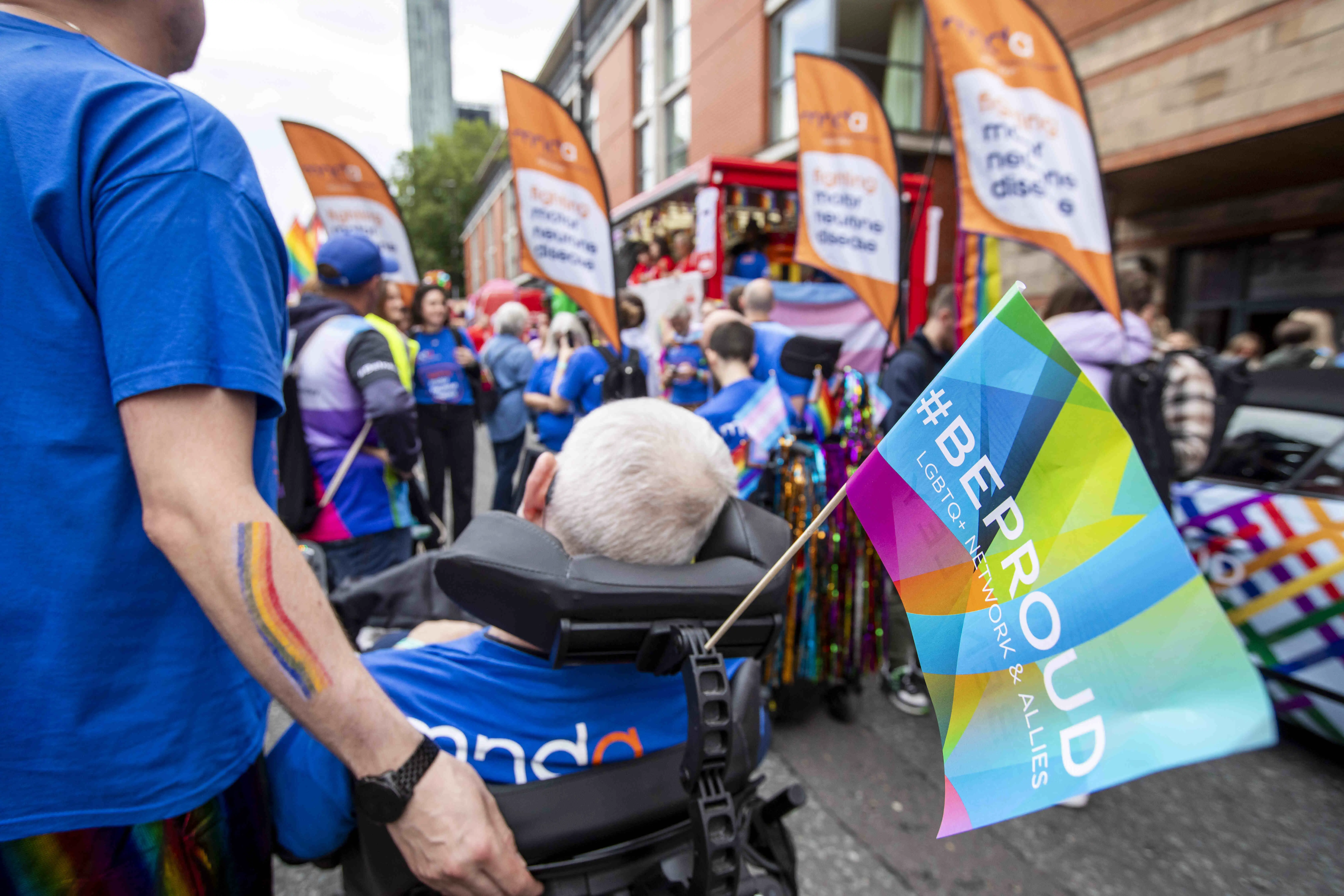Person living with MND in a mobility scooter looks on at a crowd wearing MND Association blue shirts at Manchester Pride. Association banners are visible.