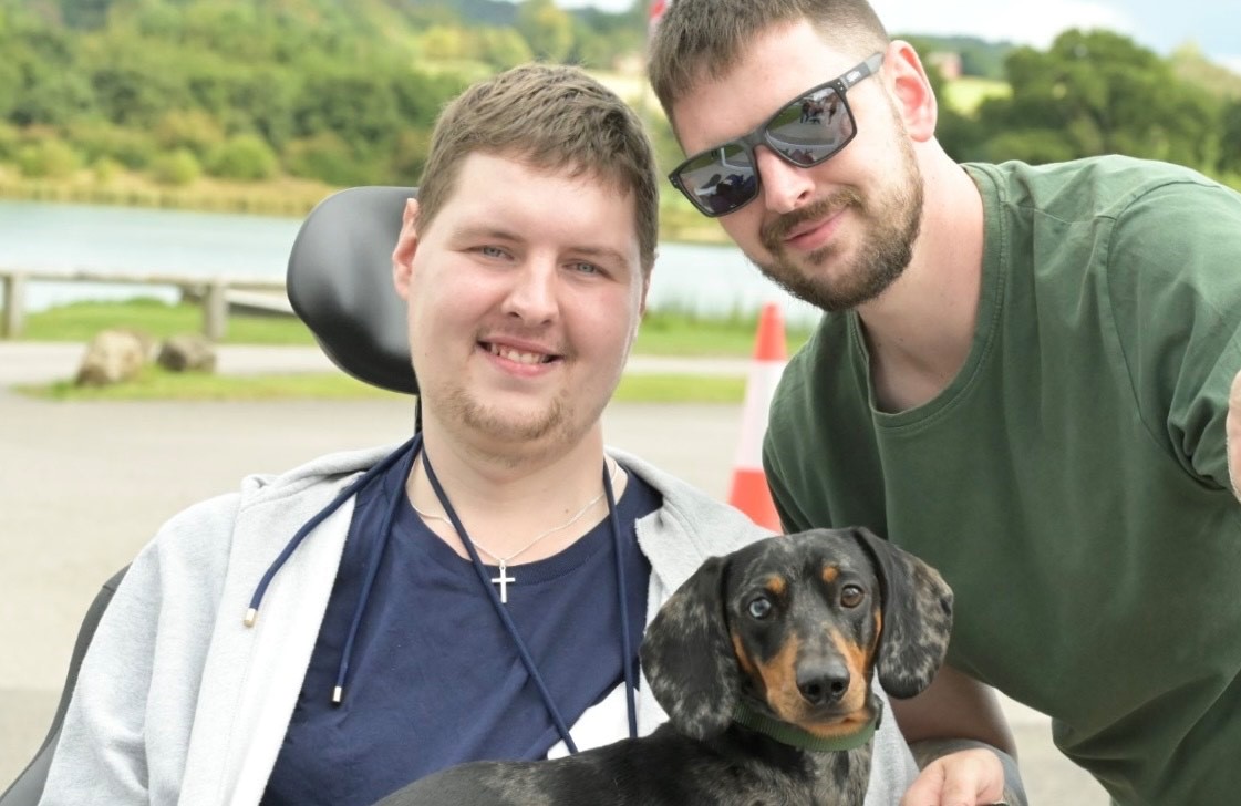 Tom with his brother Rob. Rob hits in his power wheelchair with his sausage dog on his lap.
