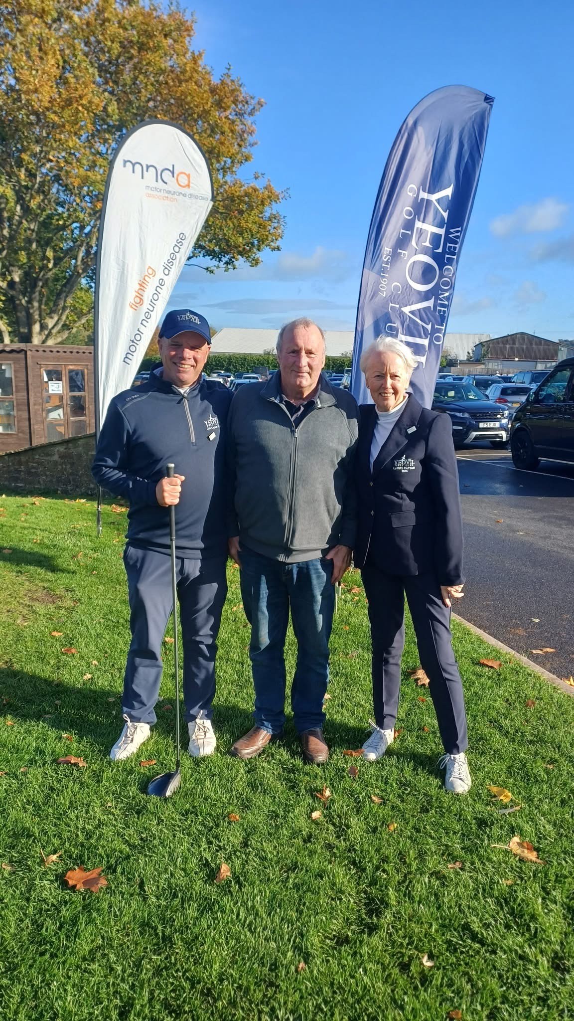 Three people including Dave Park, a person living with MND, stood in front of two flags on a grassy verge