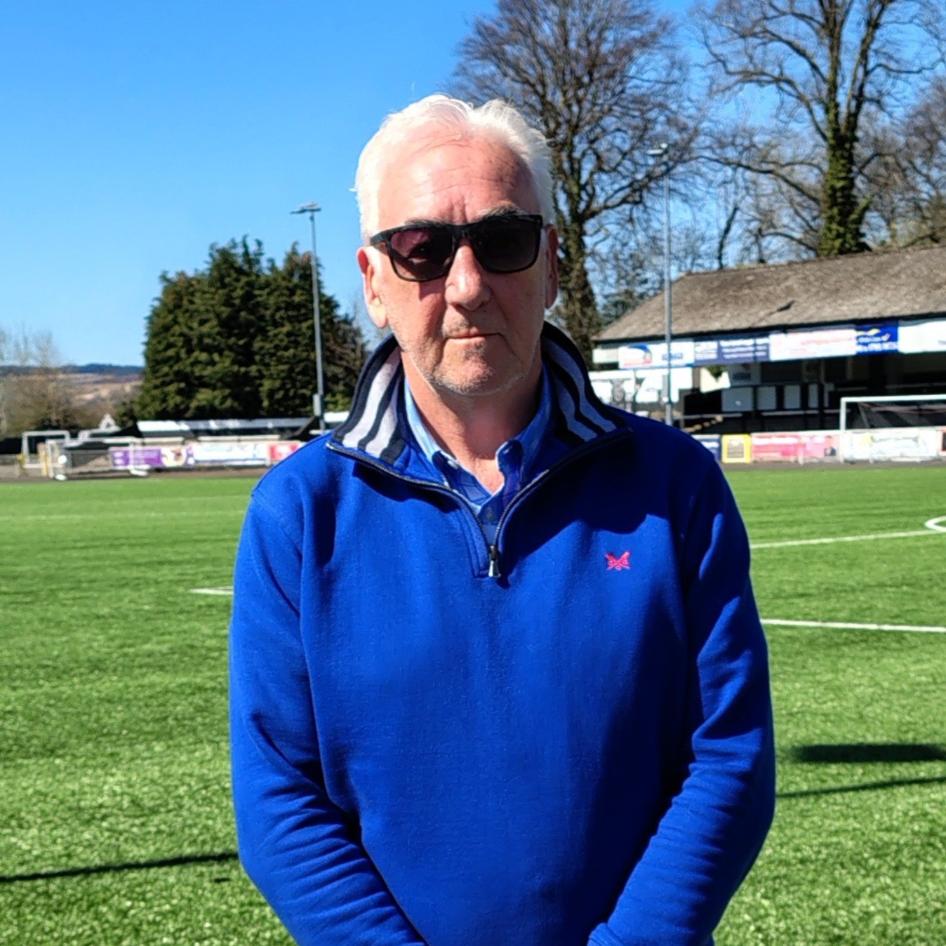 Kevin is standing on an outdoors football pitch, wearing a sunglasses and blue top and smiling at the camera.