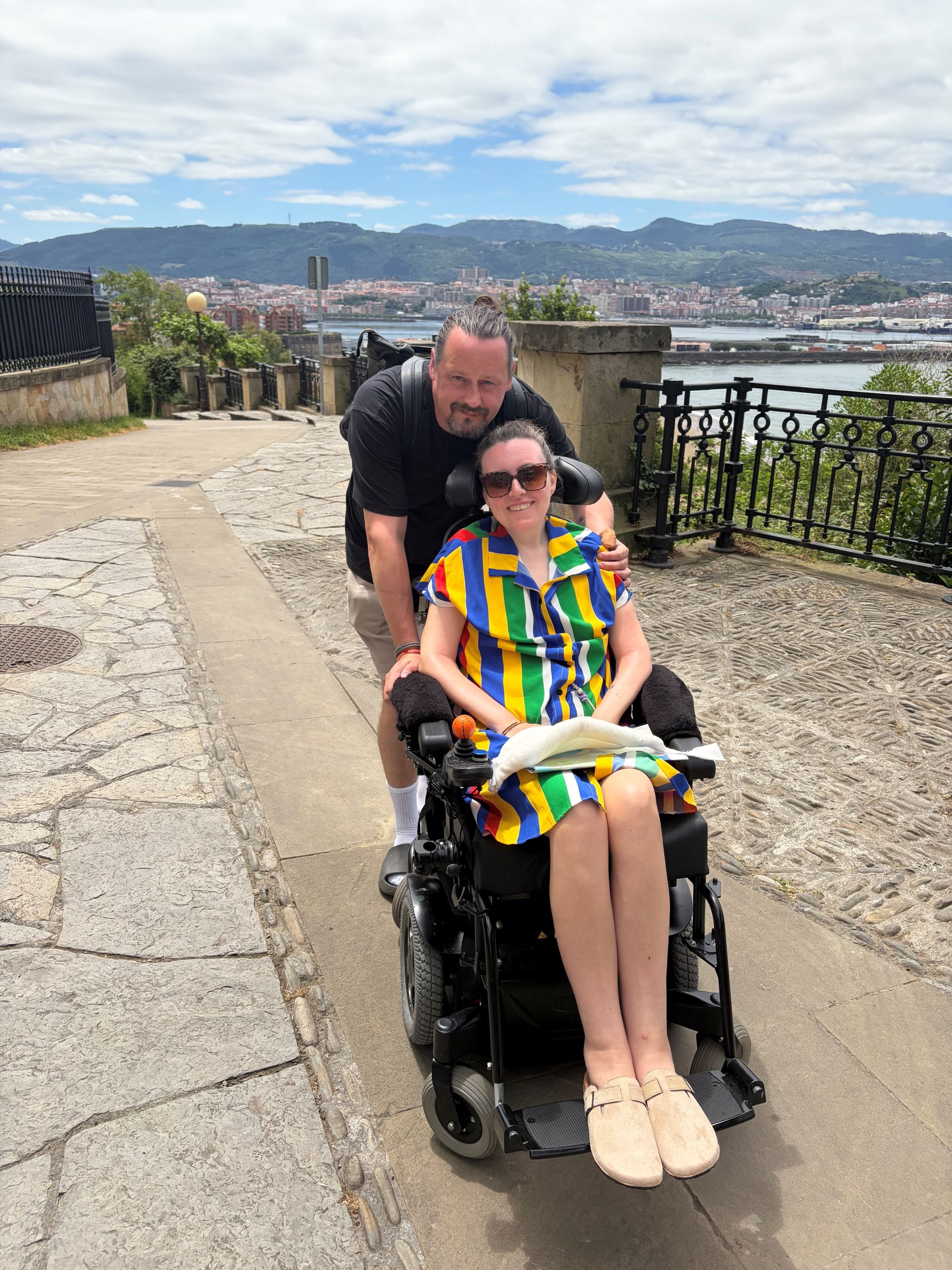 Anna and Martyn on holiday. Anna sits in her electric wheelchair, Martyn stands behind. They both smile to the camera with a panoramic view of the city behind.