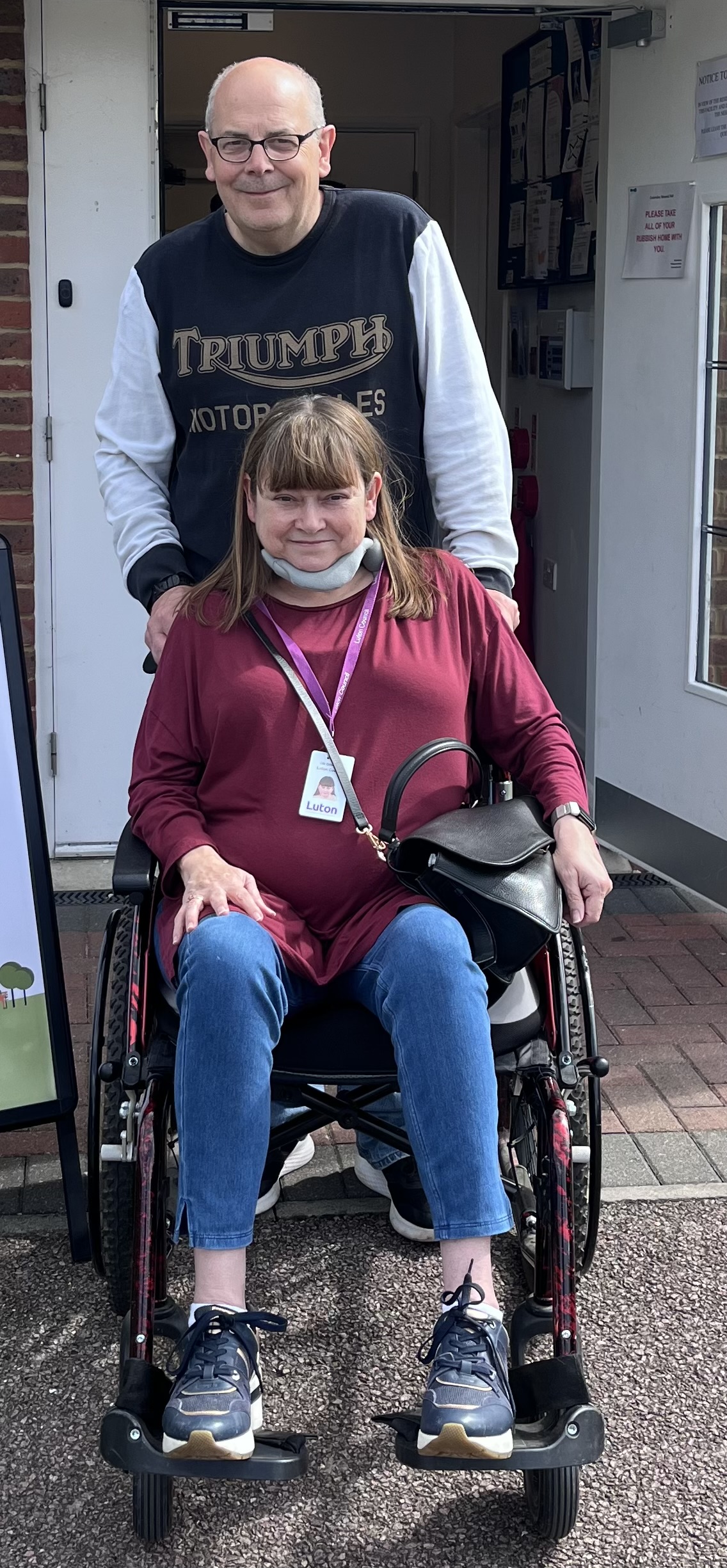 Gill in her chair with Matt standing behind her. They both smile to the camera.