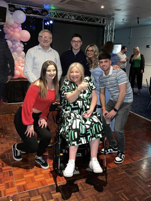 Julie and family at a party. Julie sits in her wheelchair, thumb up, smiling