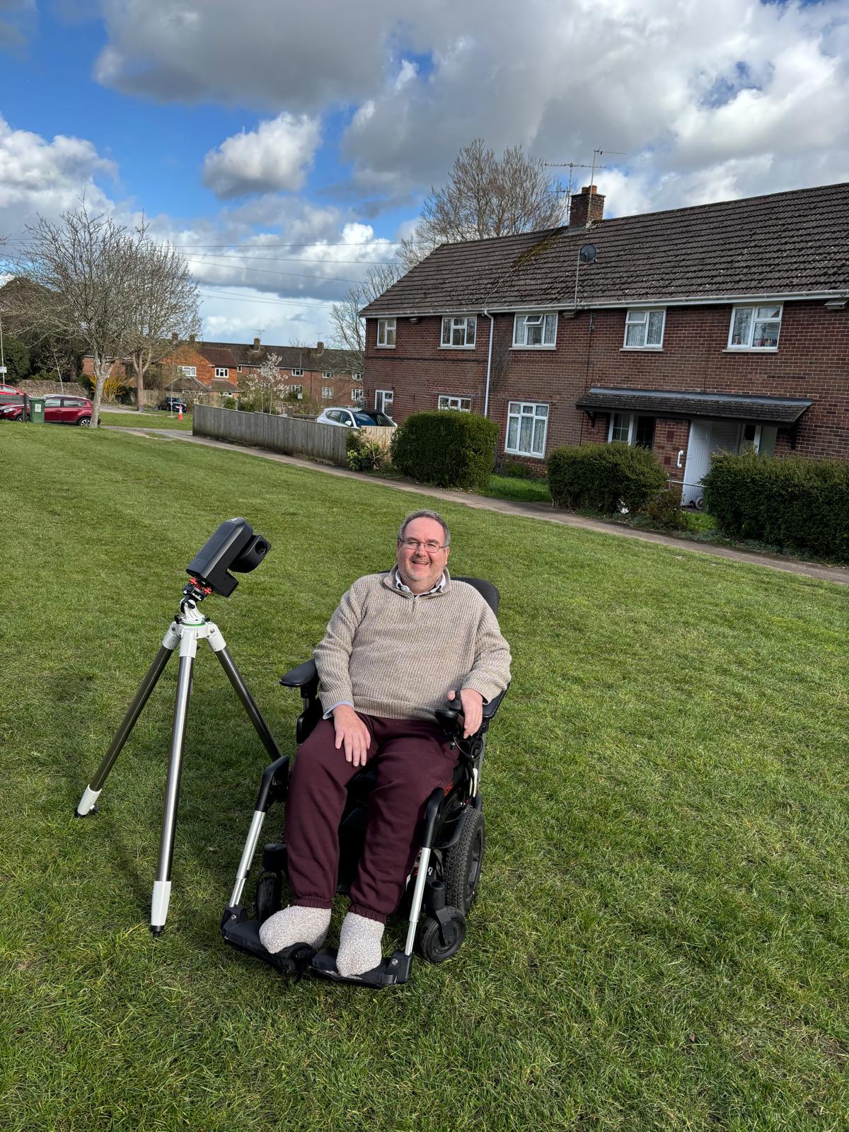 Jim sits in his wheelchair on the front green outside his house. His adapted telescope is next to him. He smiles brightly.