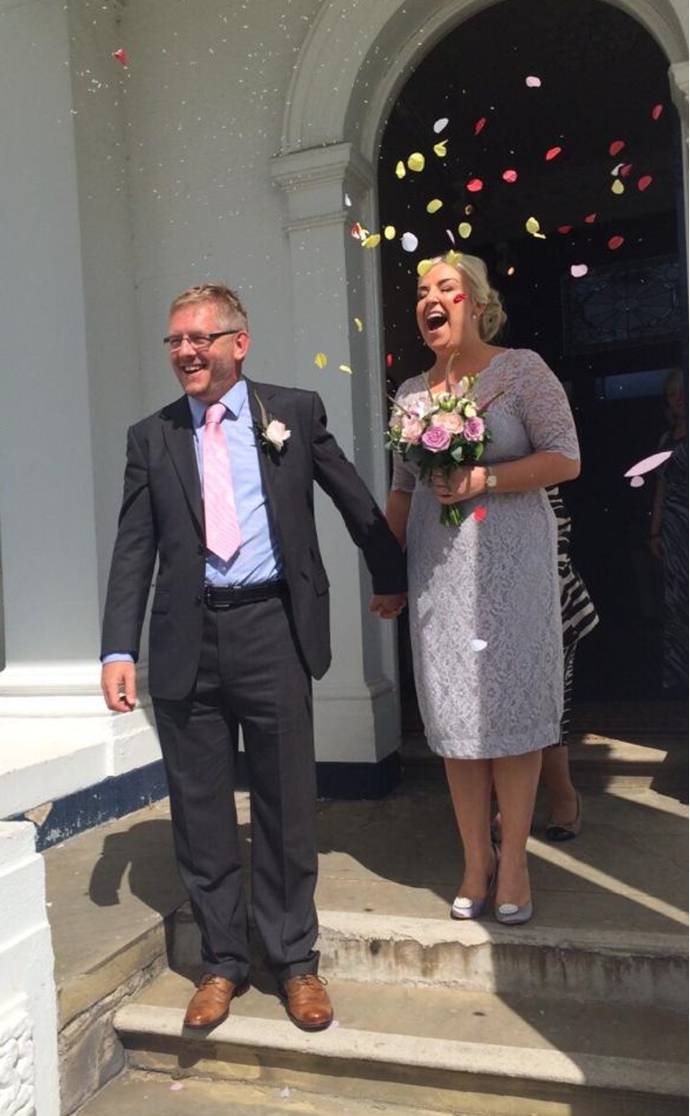 John and Julie on their wedding day. They stand at the entrance to the church, confetti in the air. They hold hands and both smile.