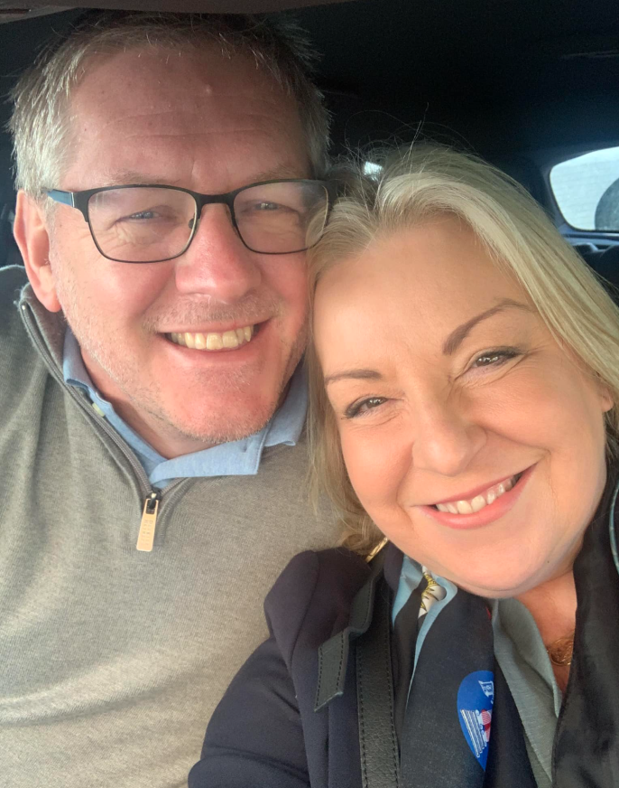 John and Julie take a selfie in the car, they both smile to the camera