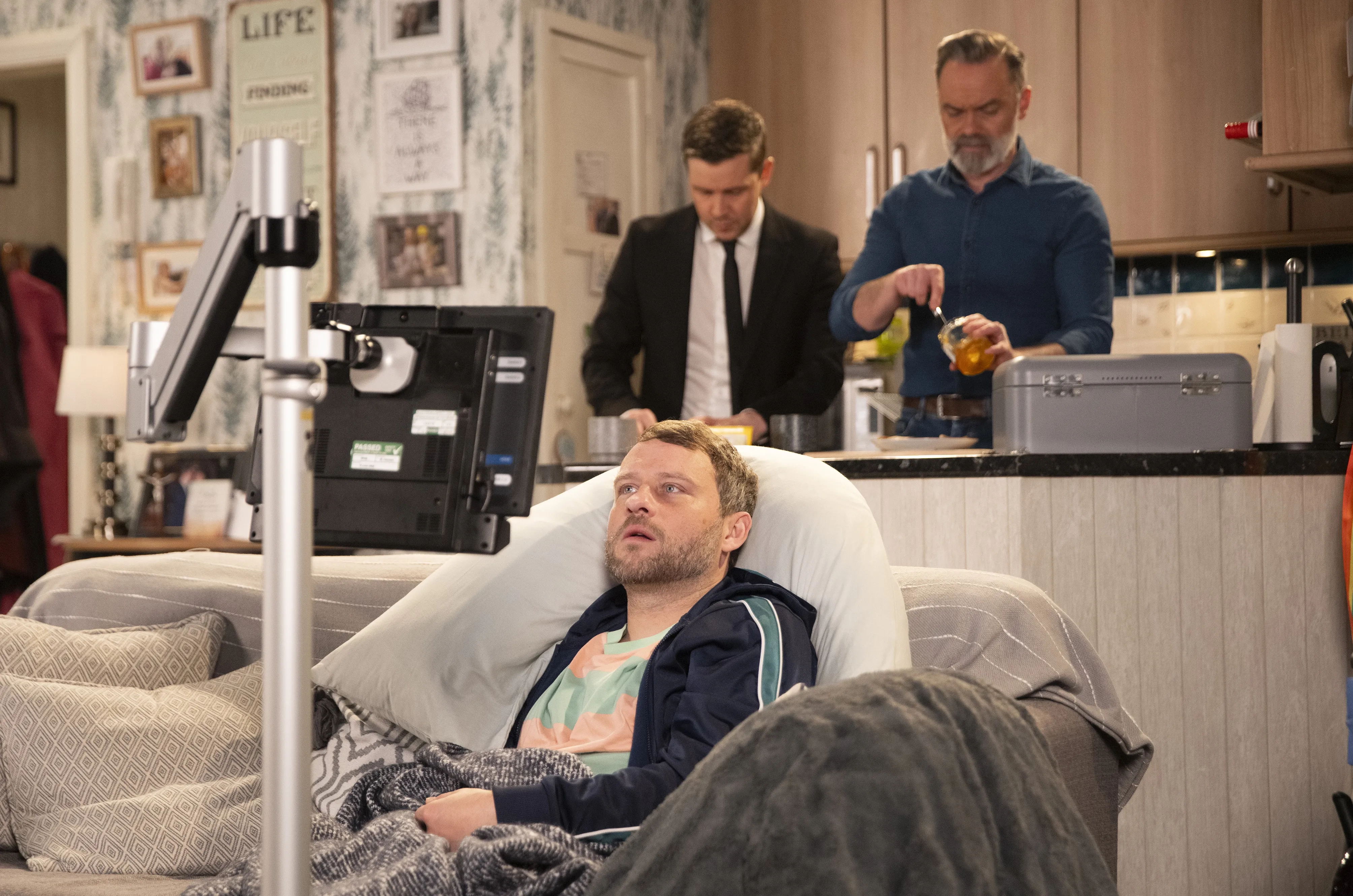Paul is in the foreground of the shot restricted to the sofa looking at his communication aid screen. Billy and Todd are in the kitchen in the background making food. 