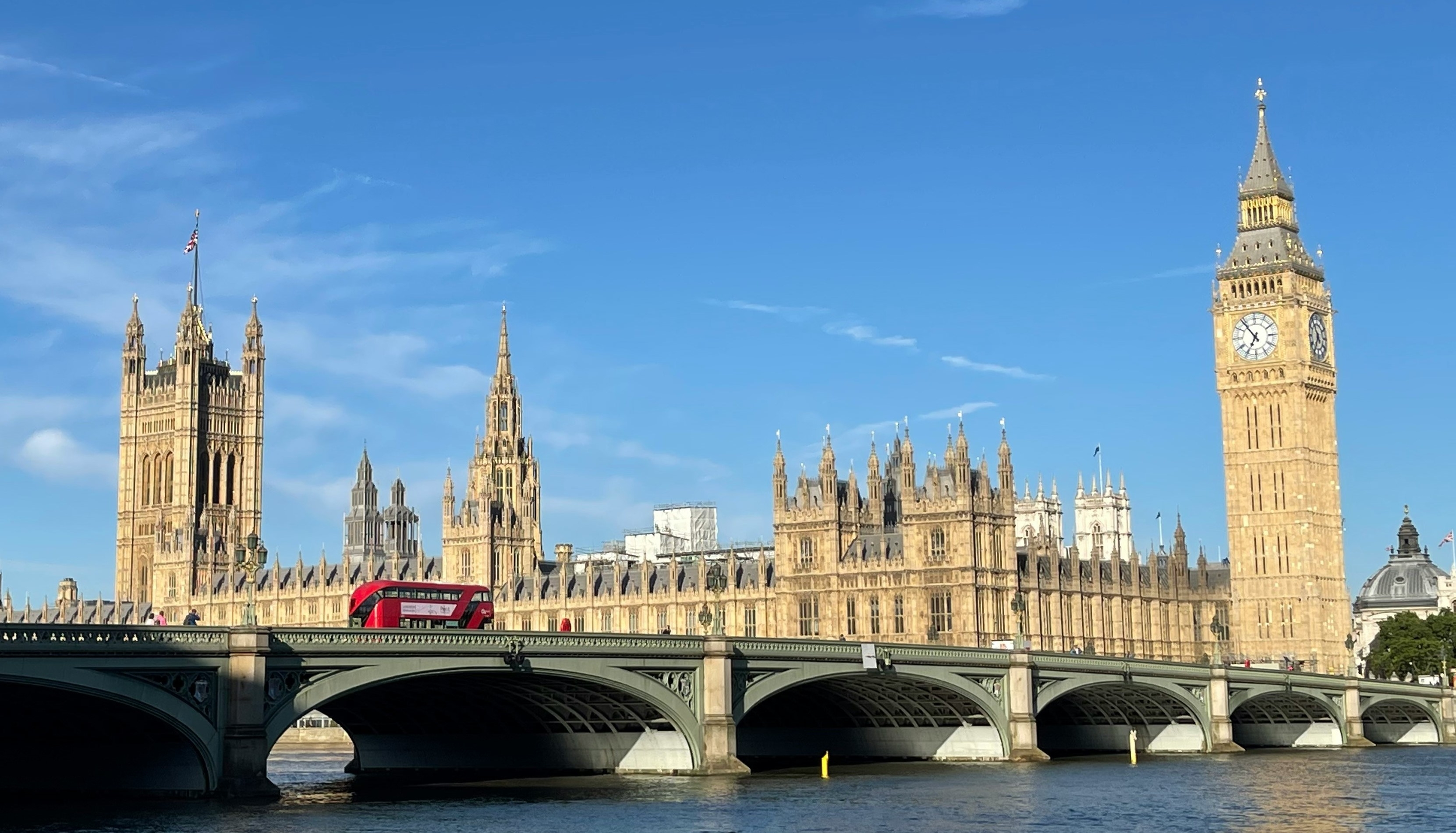 The Palace of Westminster seen from the other side of the River Thames. The sky is blue and there is a red double-decker bus crossing the bridge.