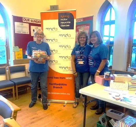 Helen Ruggles, Christine Charter and Debbie Newbound stood together in front of a MND Association banner following a bake sale fundraiser