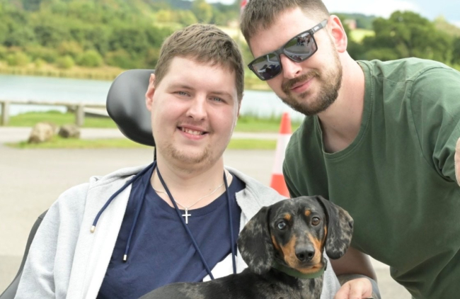 Tom with his brother Rob. Rob hits in his power wheelchair with his sausage dog on his lap.