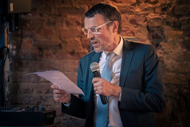 Andrew delivering his best man speech at his brothers wedding. He holds a microphone in one hand and his written speech in another. He stands in front of an exposed brick wall.