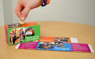Hand placing money in a fundraising box, with other fundraising materials also visible.