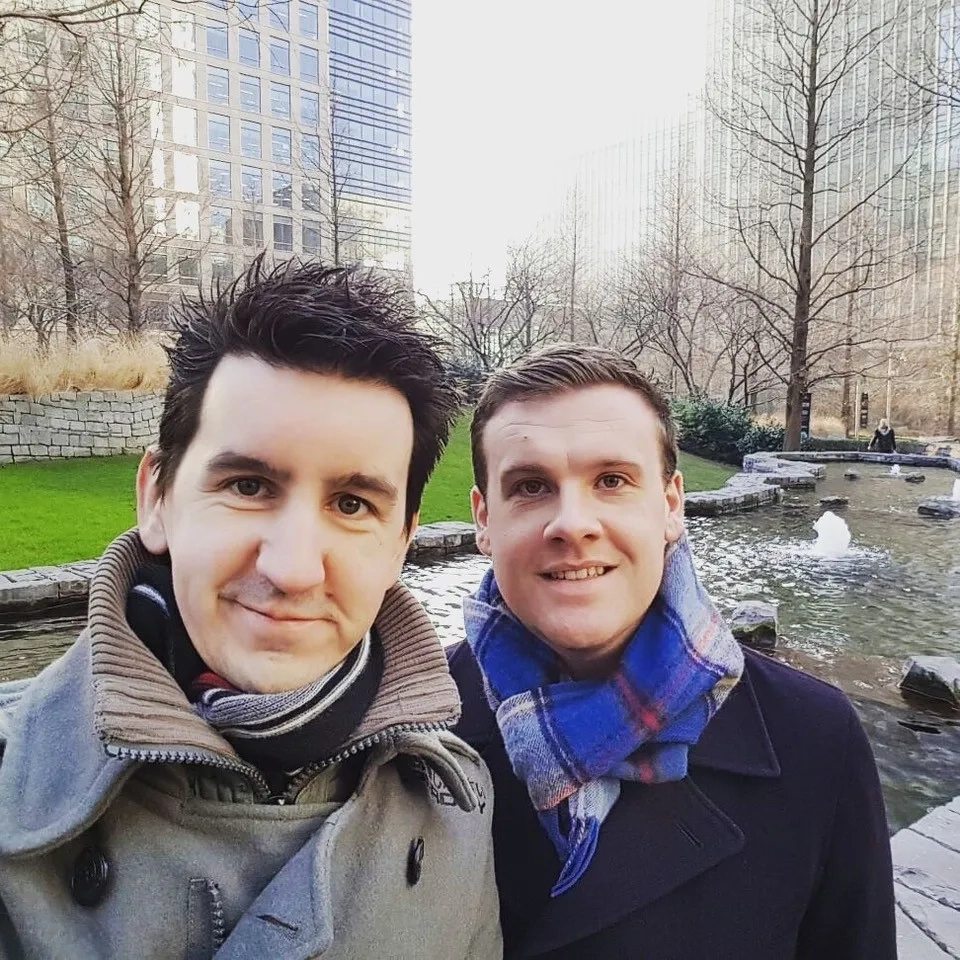 Sam and James taking a selfie outdoors. They are wearing coats. There are tall buildings and a stream or pond behind them.