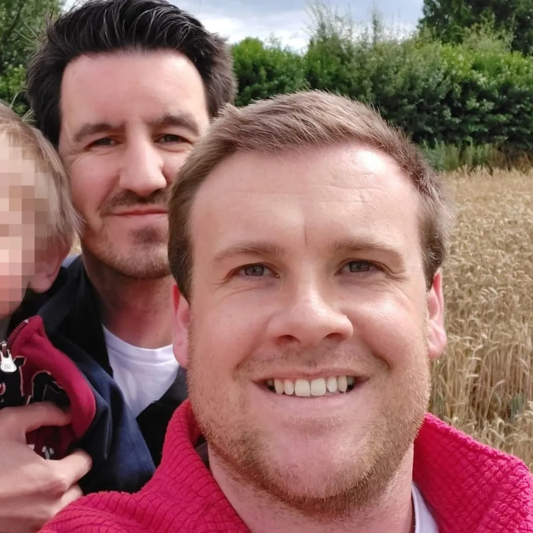 Sam and his partner taking a selfie. They are standing in a field, with their son.