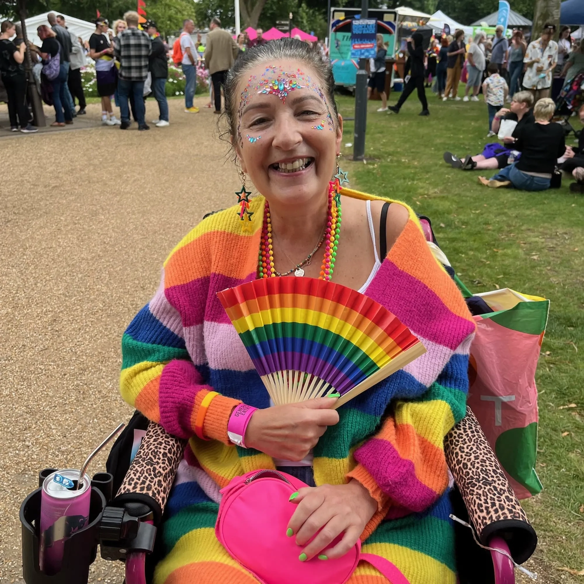 Debbie at a pride event. She is in her pink wheelchair with leopard print arm covers, in a rainbow striped outfit with a rainbow fan.