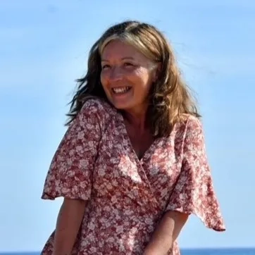 Debbie on the beach in a red floral print dress. She is smiling with the blue sky and sea behind her.
