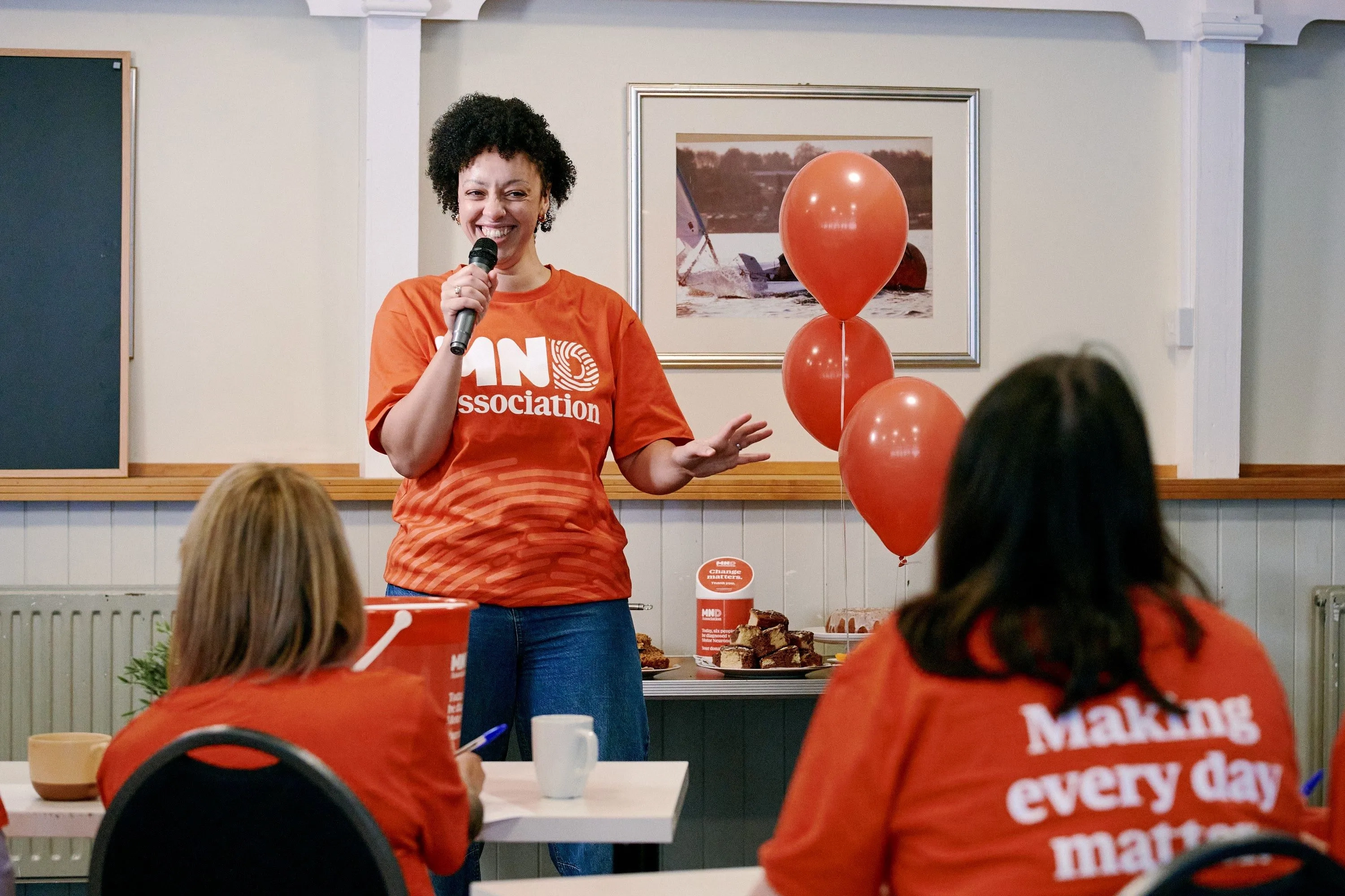 someone wearing a MND Association tshirt, speaking into the microphone in front of two people sat down
