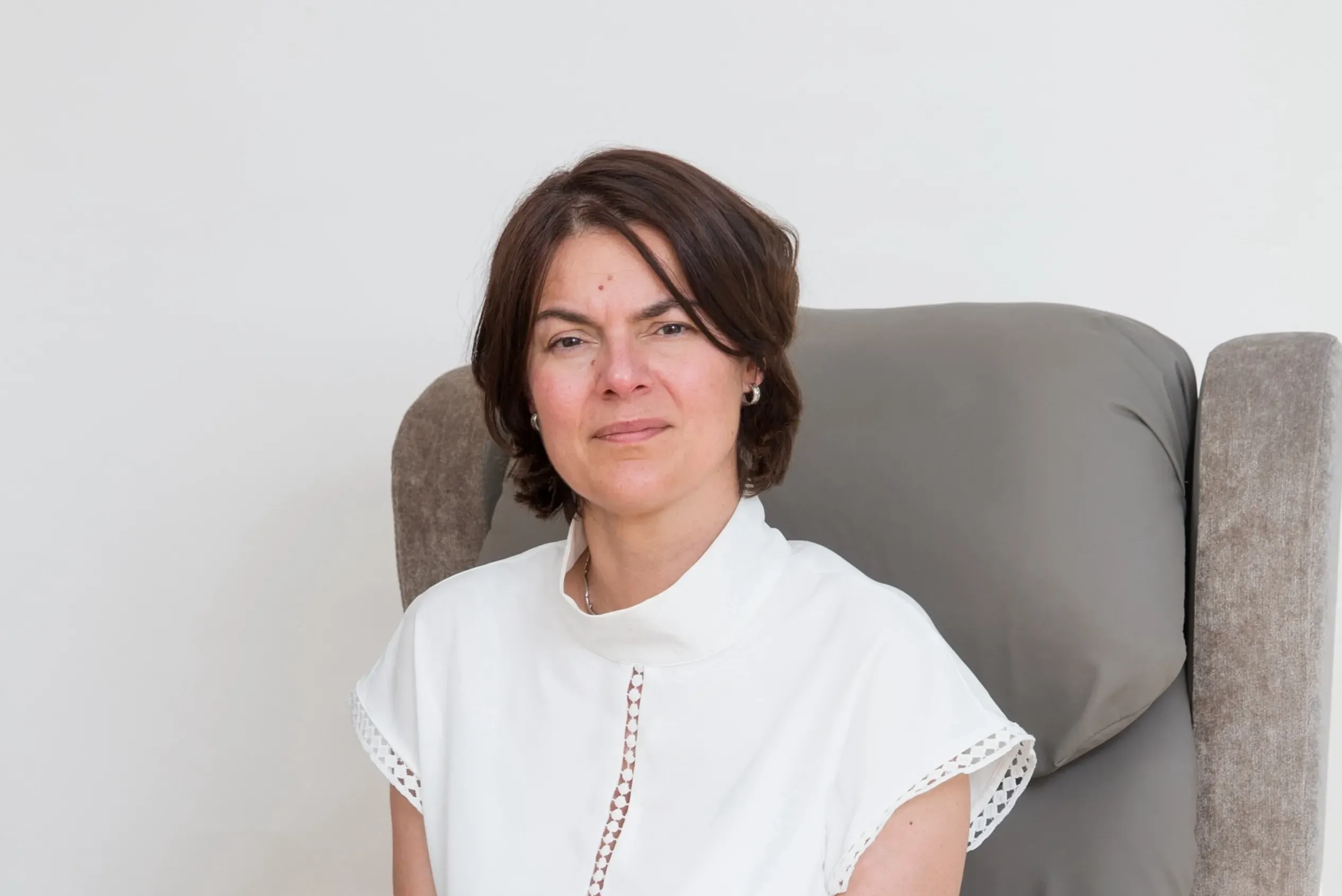 Seckin McGuirk, a person with SOD1 MND, is looking straight at the camera. She is wearing a white shirt and sitting in a grey chair.